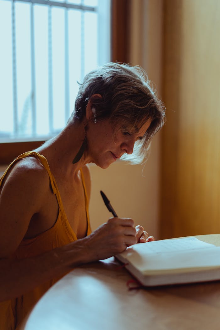 Elderly woman in a yellow top writing in a notebook at a table indoors.