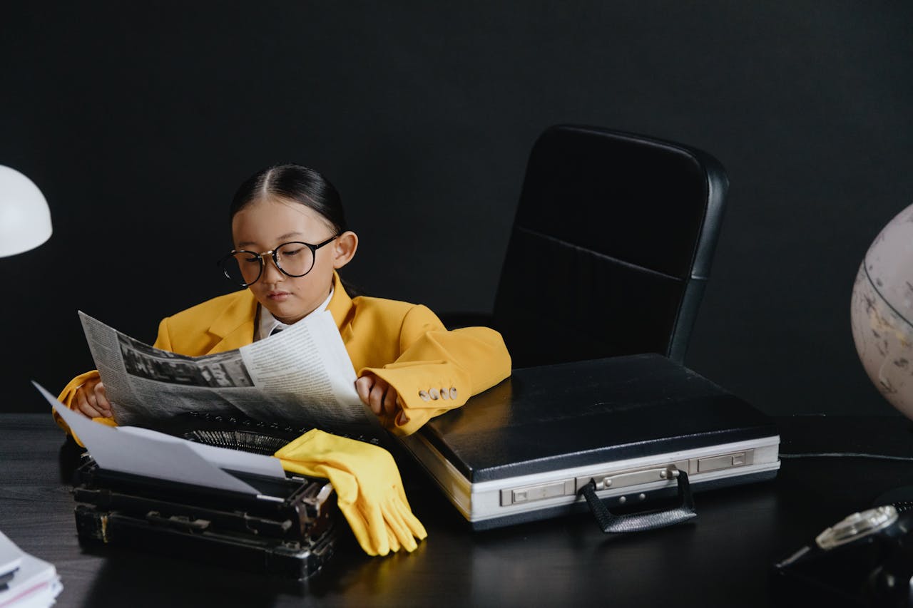 A child in eyeglasses and a yellow suit reads a newspaper at a desk, embodying a young journalist.